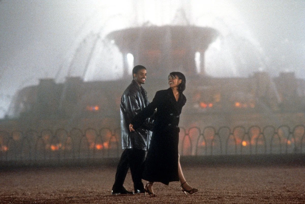 Cena do filme Love Jones. Na imagem temos a imagem de Nina (Nia Long), uma mulher negra, de cabelo curto liso preto. Ao seu lado está Darius (Larenz Tate), homem negro, cabelo preto raspado. Eles estão andando em um parque á noite, perto de uma fonte