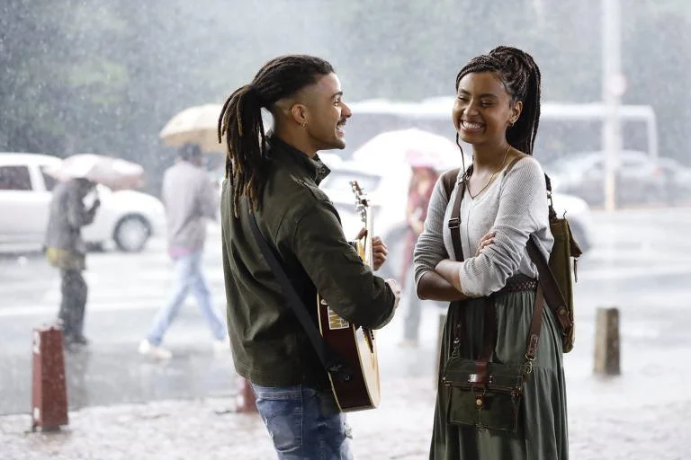 Cena do filme Um ano Inesquecível - Outono. Na imagem temos João Paulo (Lucas Leto), um jovem negro, com locs, segurando um violão. Ao seu lado está Anna Júlia (Gabz), uma jovem negra, com locs braids. Eles estão no centro da cidade, tomando chuva.