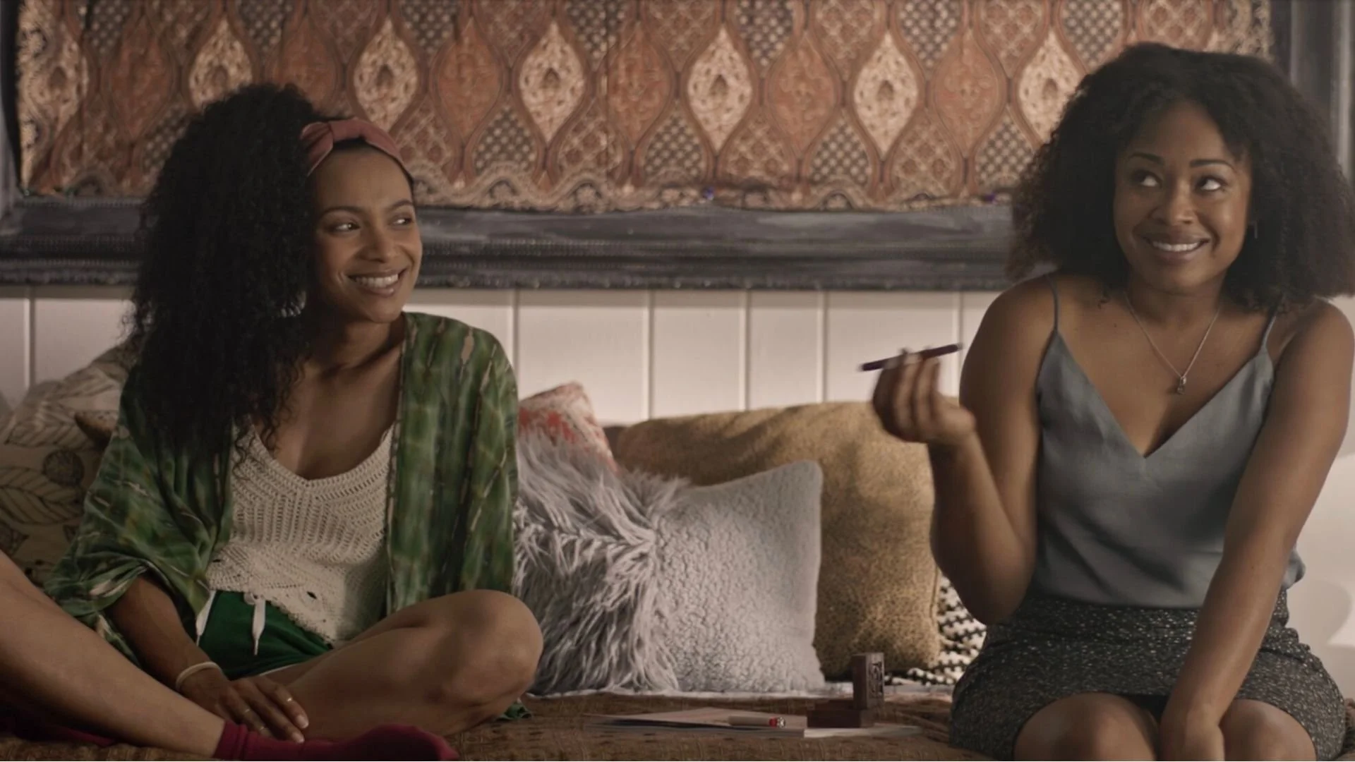 Cena do filme Tender. Na direita temos Kiara (Farelle Walker), uma mulher negra de cabelo cacheado. Ao seu lado está Lulu (Trishauna Clarke), outra mulher negra de cabelo cacheado. Elas estão sentadas em um sofá e rindo.