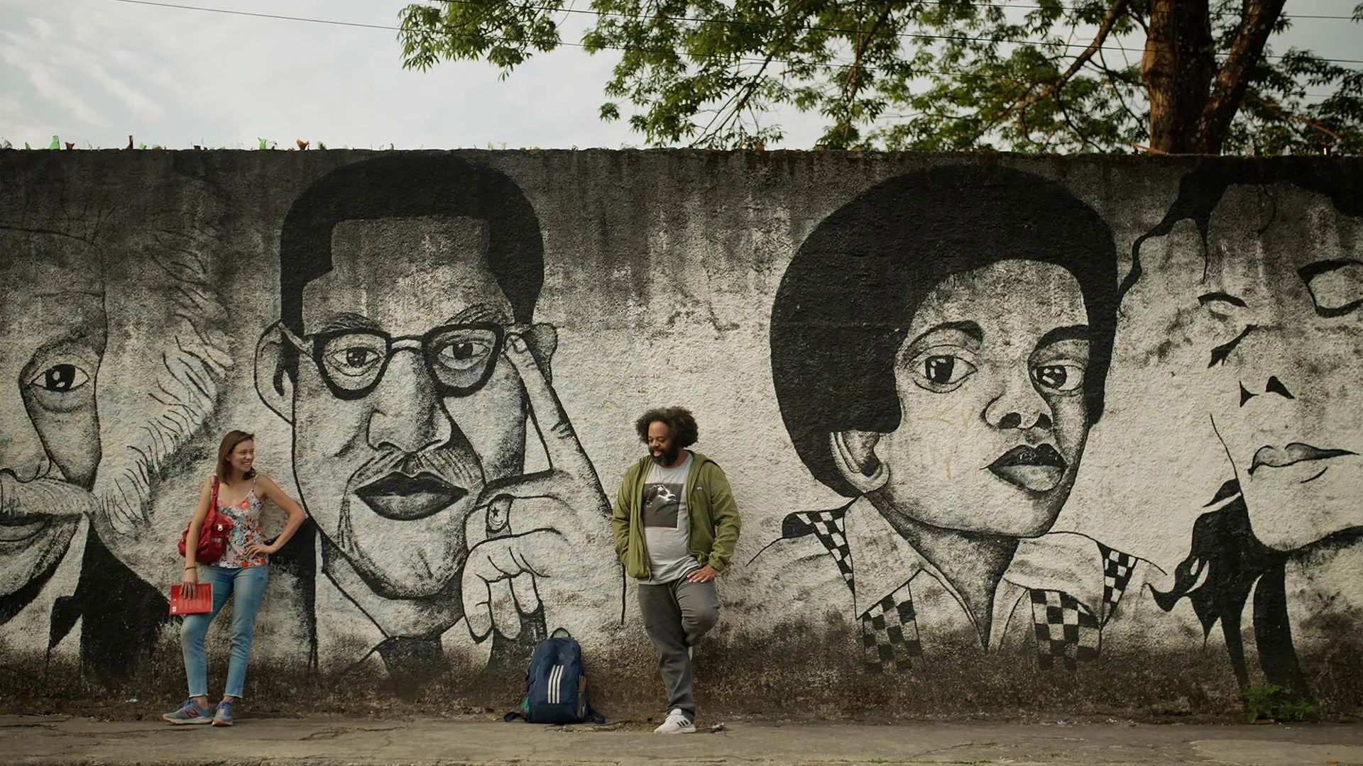 Cena do filme O Dia que te Conheci. Na  imagem temos Zeca (Renato Novaes), homem negro, com cabelo black. Ao fundo tem um mural com a arte de grandes nomes históricos, Albert Einstein, Malcom X, Michael Jackson criança e adulto.