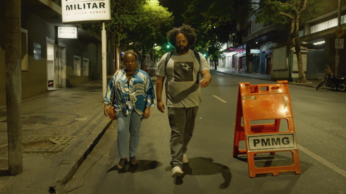 Cena do filme O Dia que te Conheci. Na esquerda temos Luísa (Grace Passô), uma mulher negra, com locs braids loiro. Ao seu lado está Zeca (Renato Novaes), homem negro, com cabelo black. Eles estão andando juntos no centro da cidade.