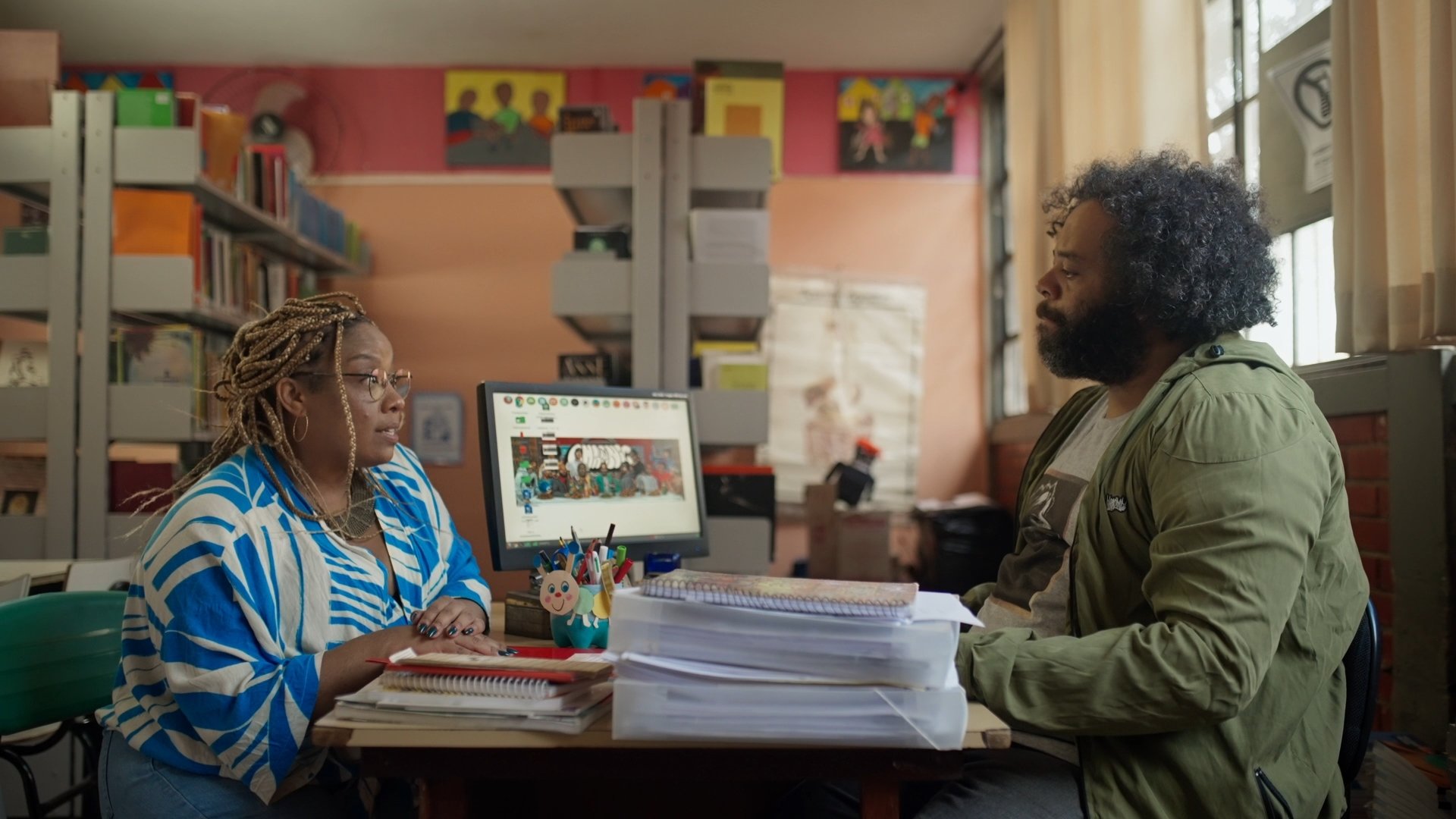 Cena do filme O Dia que te Conheci. Na esquerda temos Luísa (Grace Passô), uma mulher negra, com locs braids loiro. Ao seu lado está Zeca (Renato Novaes), homem negro, com cabelo black. Eles estão dentro de uma biblioteca escolar, se olhando.
