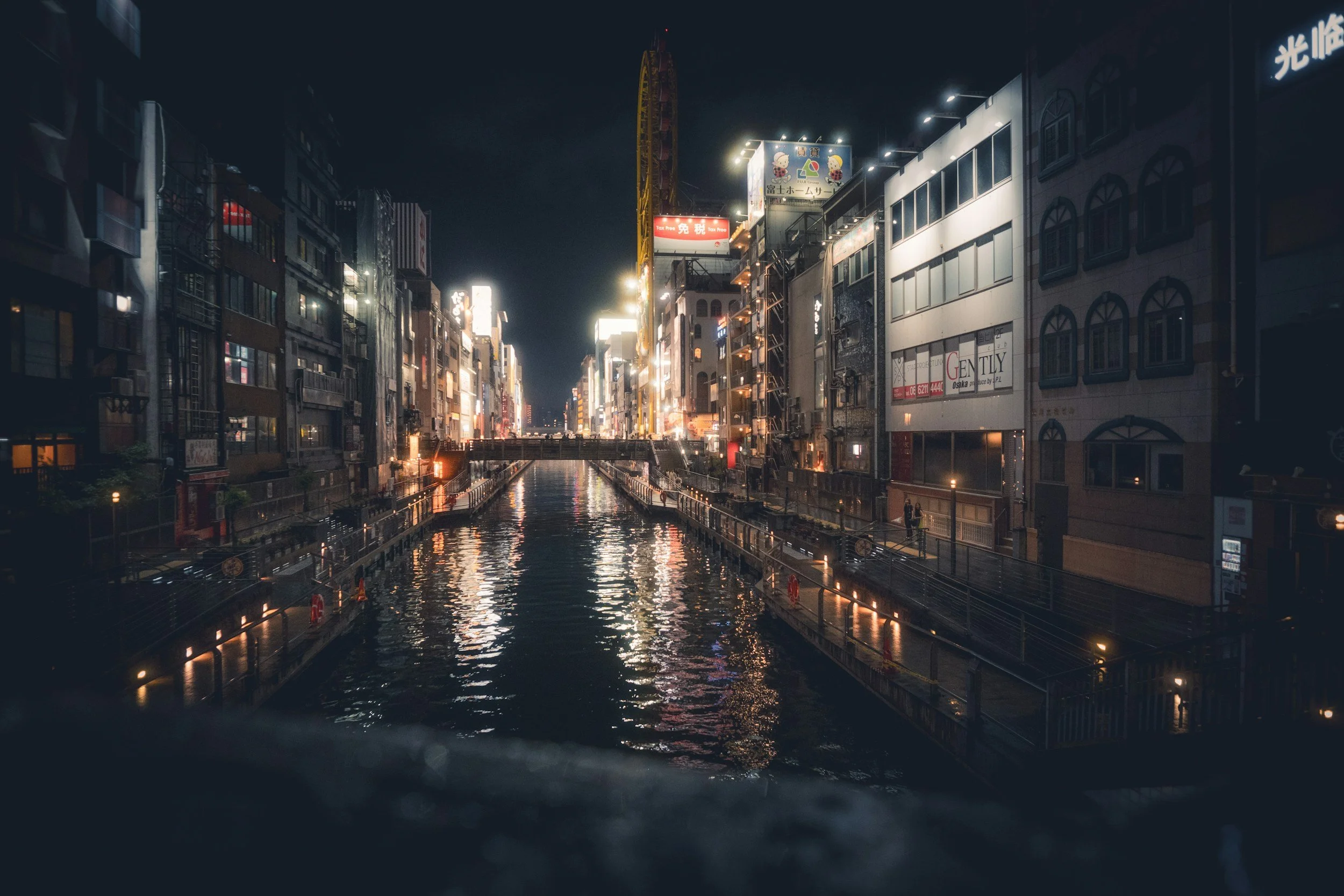 Nighttime cityscape with illuminated buildings lining a canal, reflecting lights on the water, with a bridge in the foreground.