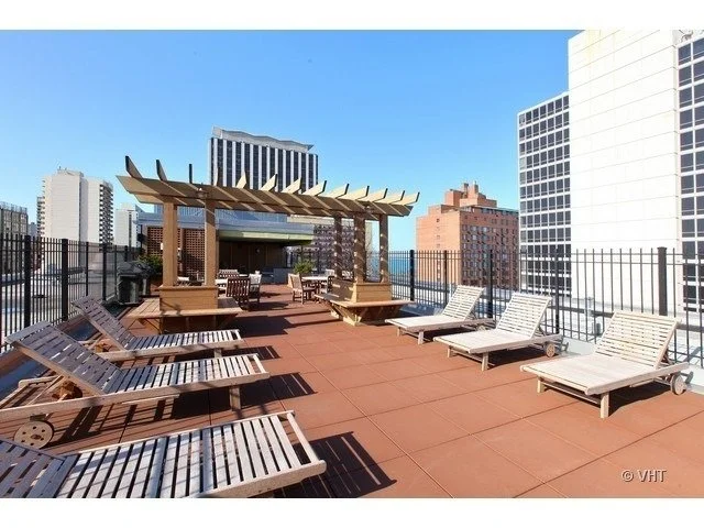 Rooftop terrace with wooden lounge chairs and a pergola, surrounded by a metal fence, overlooking city buildings and a clear blue sky.