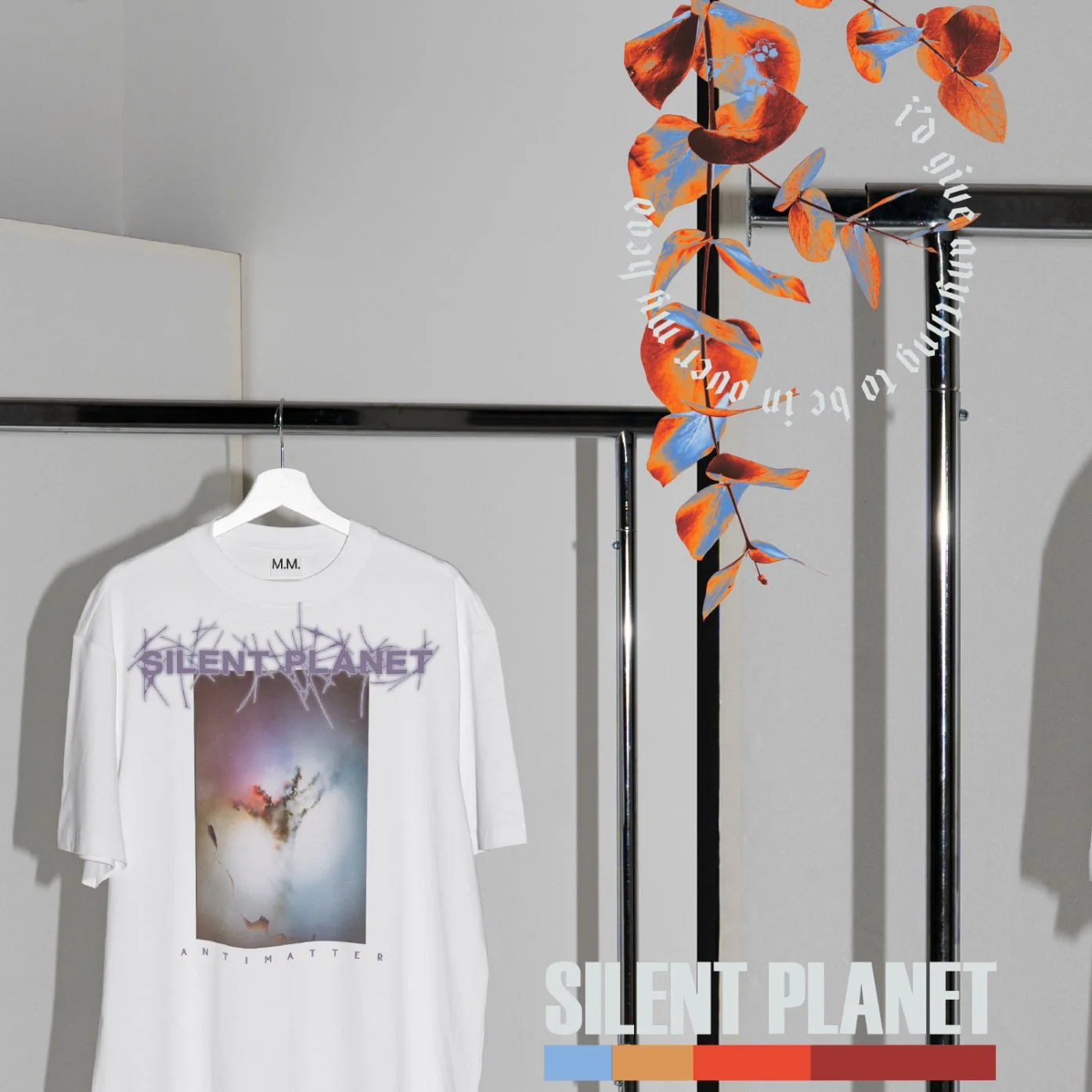 Un t-shirt blanc avec un design artistique, affichant le texte "Silent Planet" en haut et "AntiMatter" en bas, suspendu sur un portemanteau en métal. À côté, une décoration florale en feuilles orange et bleues pend le long du cadre.