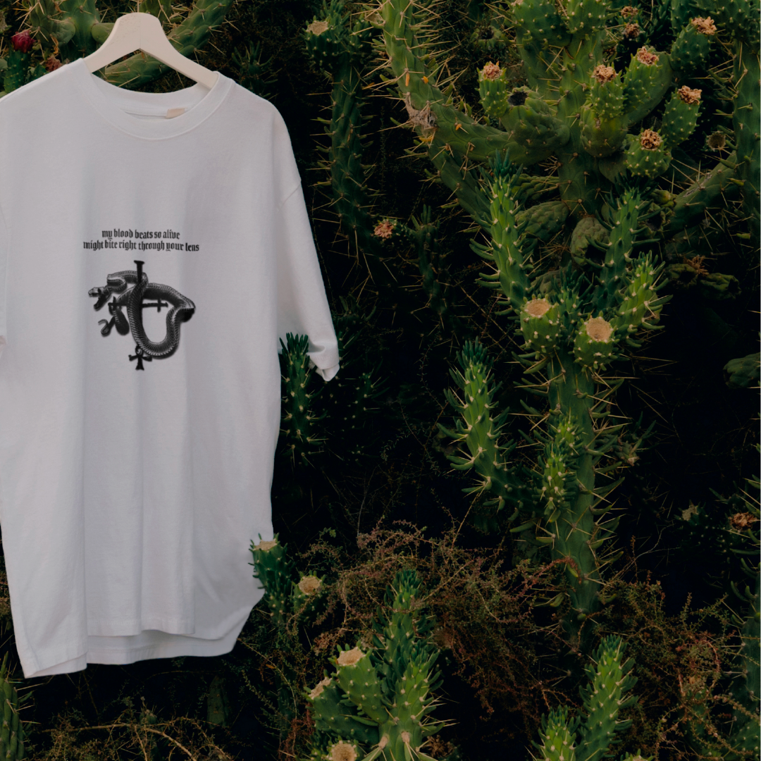 Un t-shirt blanc avec un graphique de serpent et le texte "my blood beats so alive might bite right through your lens" accroché à une branche parmi des cactus verts épineux.