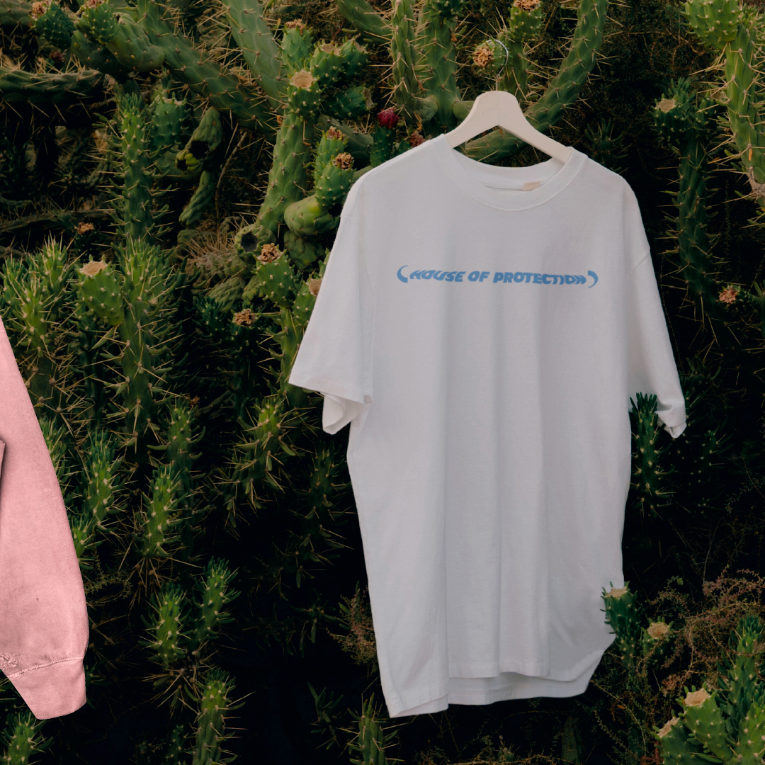 T-shirt blanc suspendu à un cintre contre un fond de cactus, avec le texte '(HOUSE OF PROTECTION)' imprimé en bleu à l'avant.