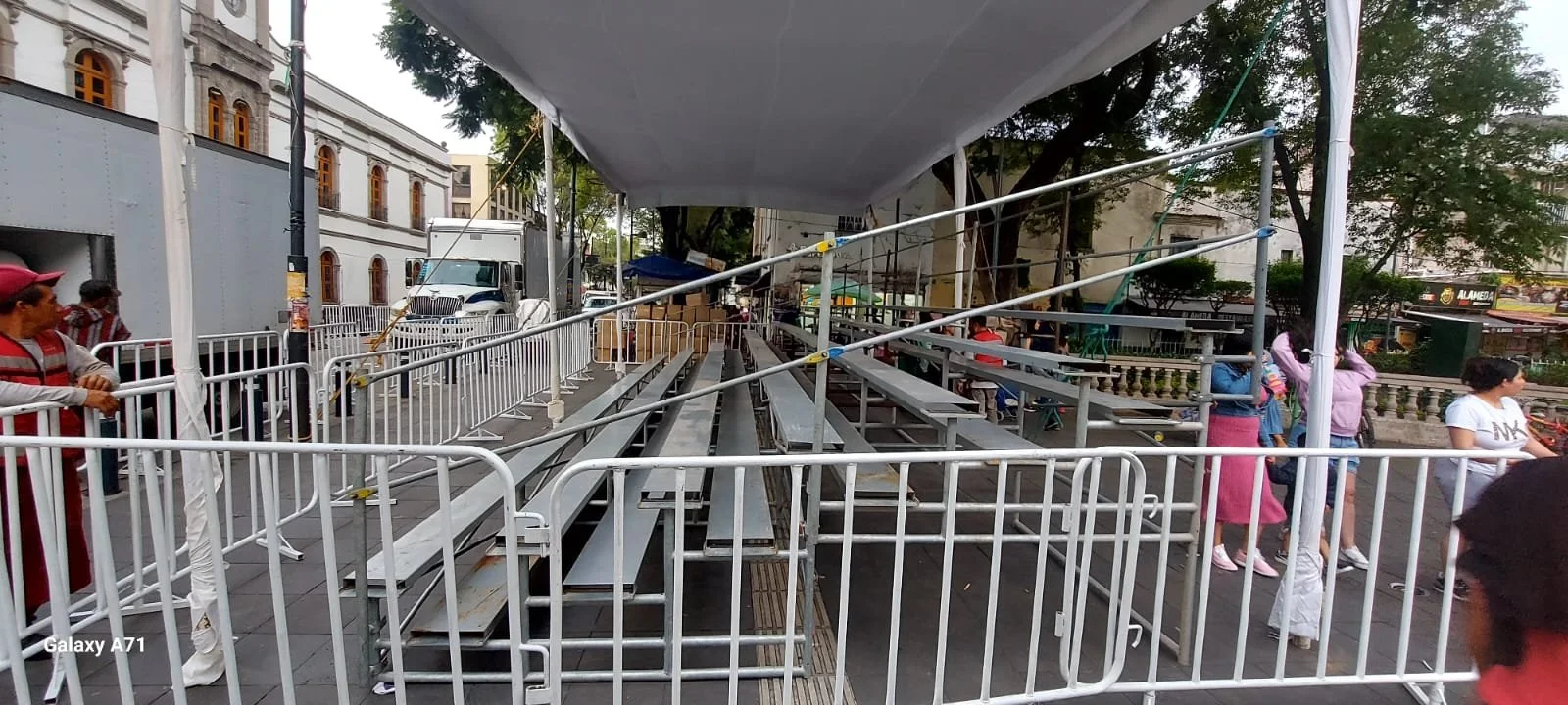 Estructura temporal con gradas de metal y rejas, aparentemente en preparación para un evento público, con varias personas alrededor en un espacio urbano.
