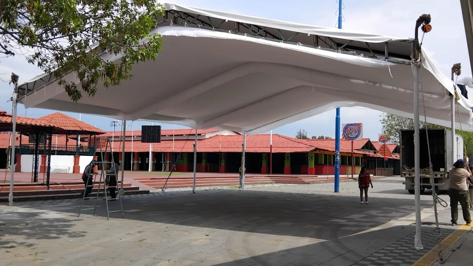 Vista de una estructura de toldo blanca en proceso de montaje en una plaza con personas trabajando y edificios de techos rojos al fondo.