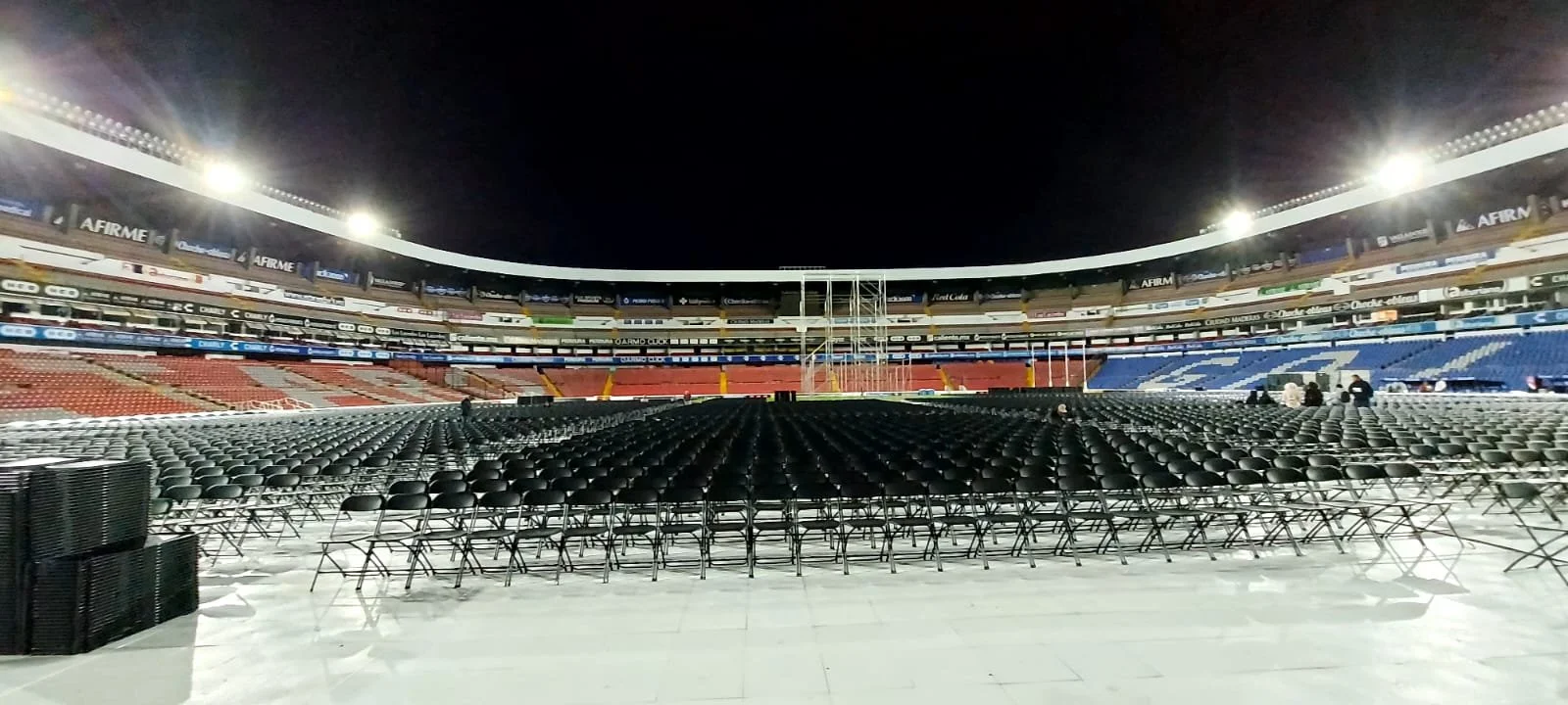 Vista de un estadio de fútbol vacío con manyas o sillas negras en el campo, iluminado por luces de noche.