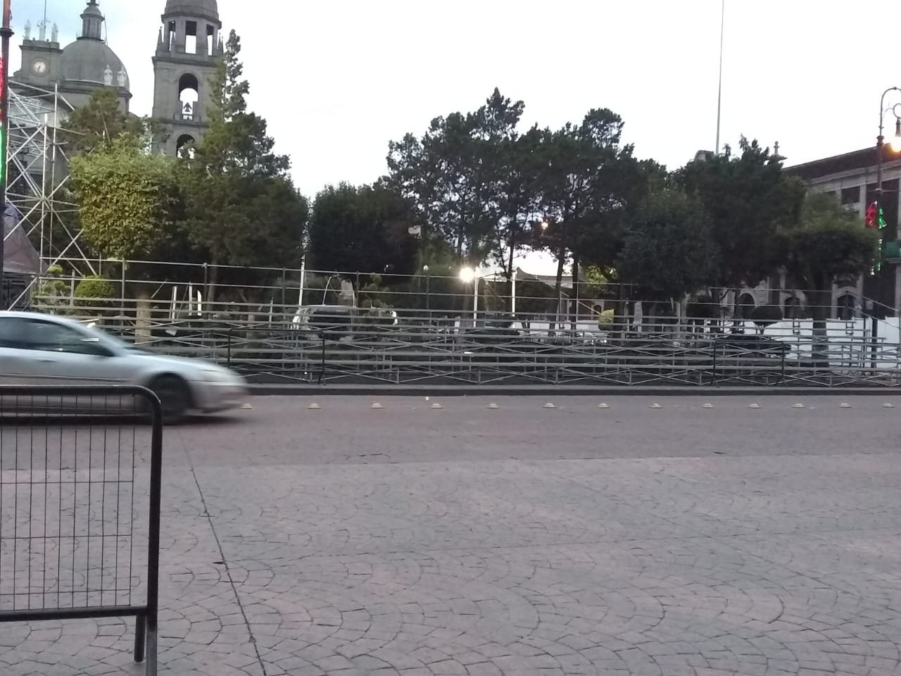 calles con edificios históricos y árboles, con una fila de vallas de metal y un coche en movimiento en primer plano.