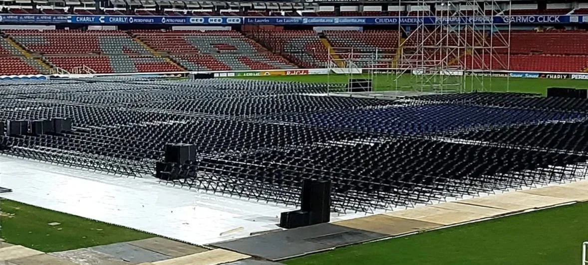 Interior de un gran evento con muchas sillas vacías, una pantalla gigante y una carpa grande de estructura metálica