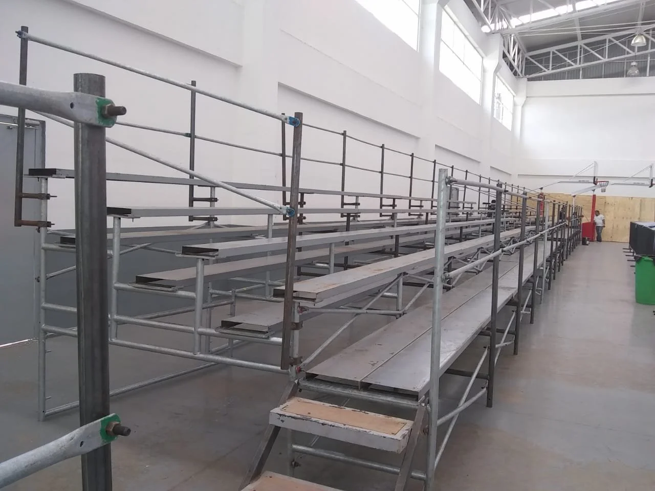 Gradas de madera y metal en un gimnasio cubierto, con una cancha de baloncesto al fondo y paredes blancas con ventanas altas.