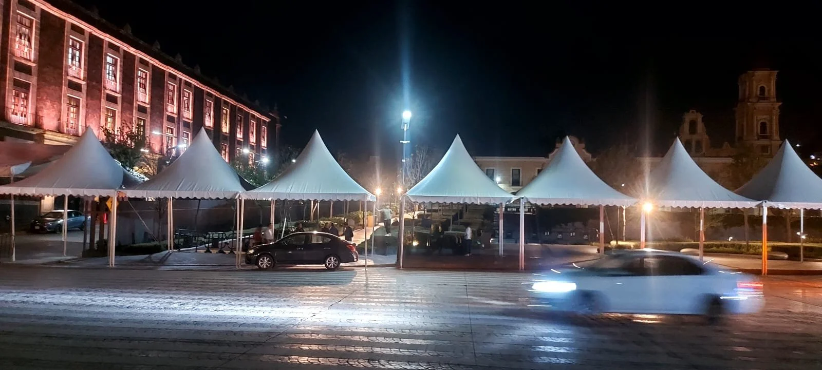 Tendas blancas en una plaza iluminada de noche, con edificios históricos al fondo y vehículos pasando por la calle.
