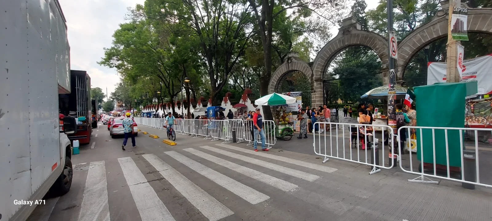 Persona caminando y bicicletas en la acera, con puestos de venta y una reja grande en la entrada de un edificio con árboles al fondo.