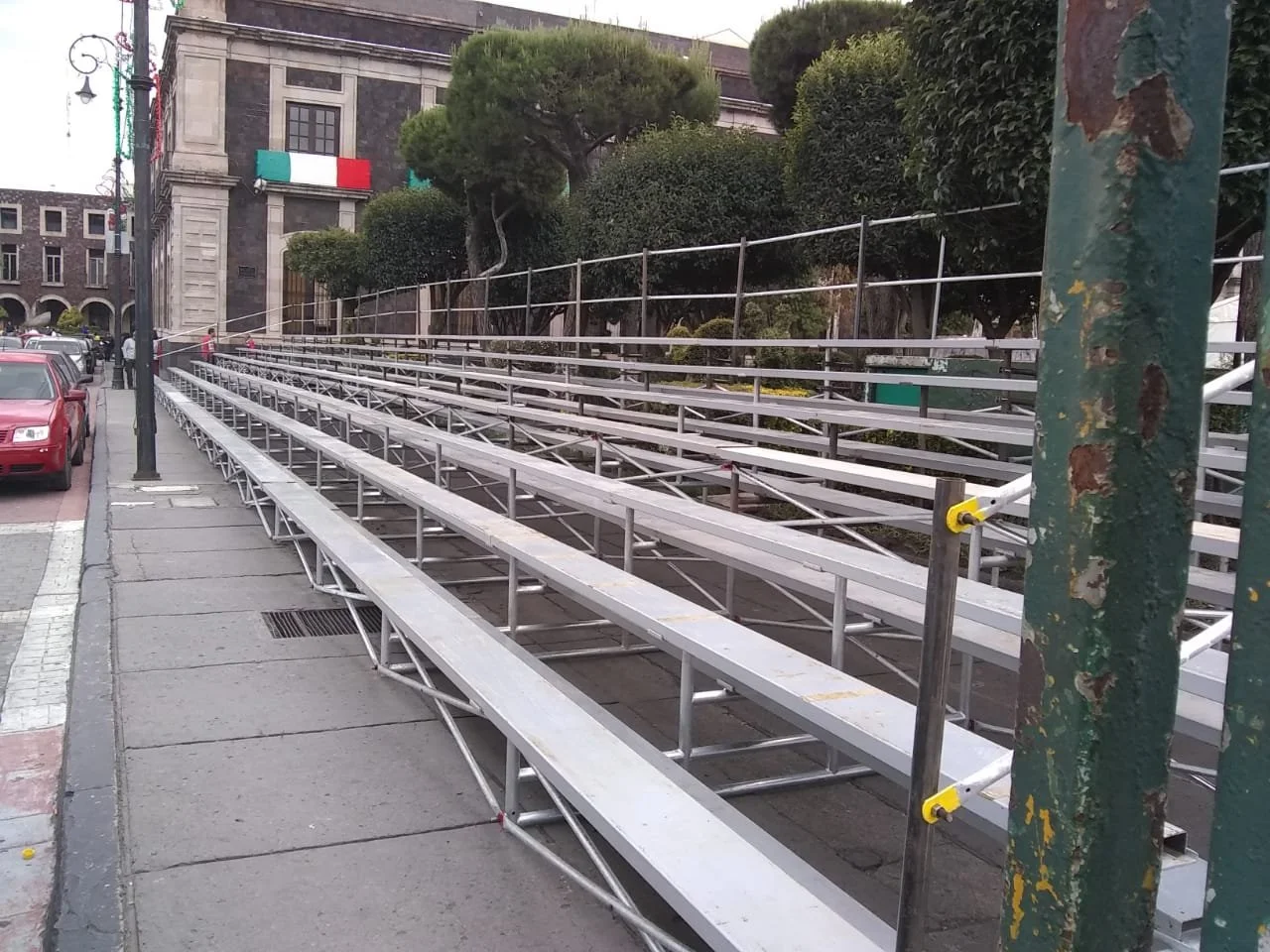 Gradas metálicas en una calle de la ciudad con árboles y un edificio al fondo que tiene banderas de México y una bandera de Italia.