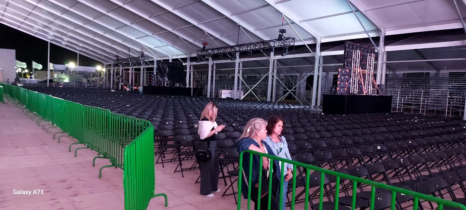 Interior de un recinto cubierto con muchas sillas negras dispuestas en filas, un escenario con iluminación y equipo de sonido, y unas pocas personas acomodándose o preparando el lugar.