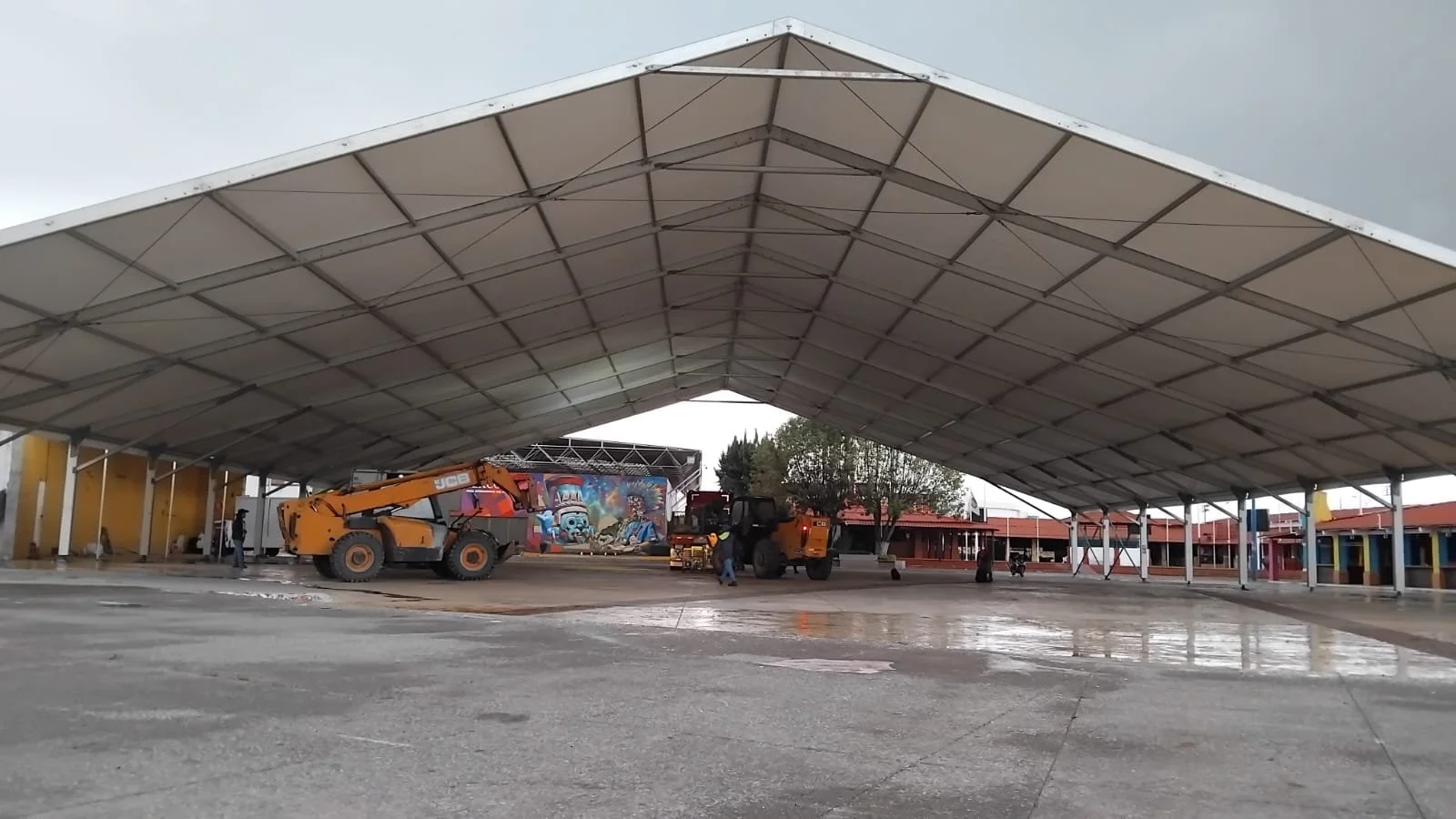 Un gran toldo de estructura metálica con maquinaria de construcción debajo, en un espacio abierto con plantas y fondo de murales coloridos.