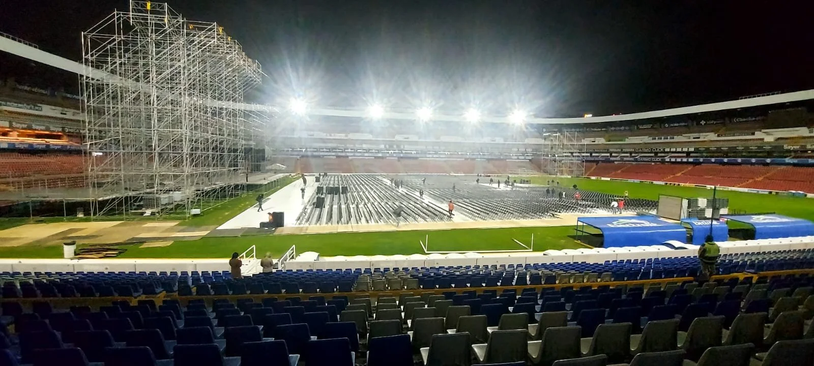 Vista del interior de un estadio de fútbol en construcción, con andamios grandes en un lado y cadenas de sillas vacías en las gradas, iluminación fuerte ilumina el campo vacío donde se están instalando asientos.
