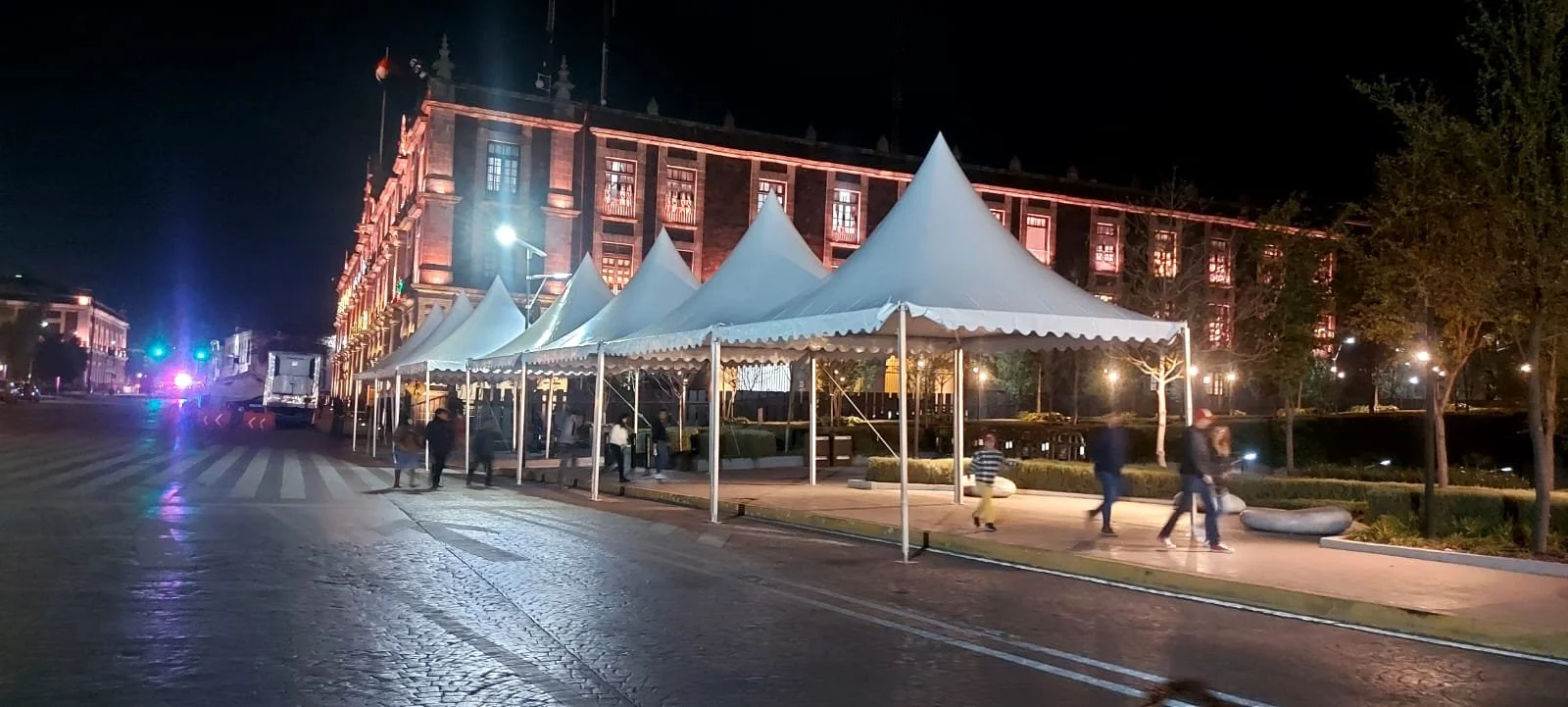 Imagen de un edificio antiguo iluminado de noche con carpas blancas en la acera y personas caminando por la calle.
