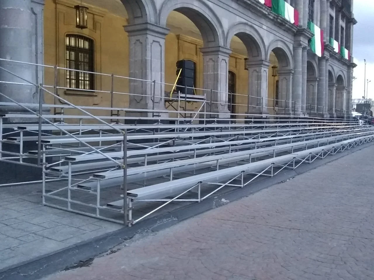 Escaleras metálicas y gradas temporales en la acera frente a un edificio con arcos y banderas mexicanas.