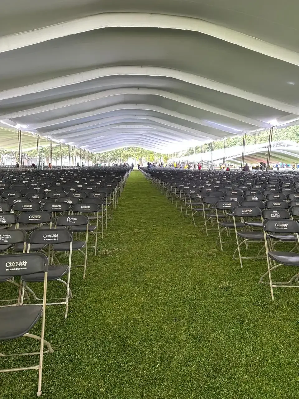 Interior de un gran evento con muchas sillas vacías, una pantalla gigante y una carpa grande de estructura metálica