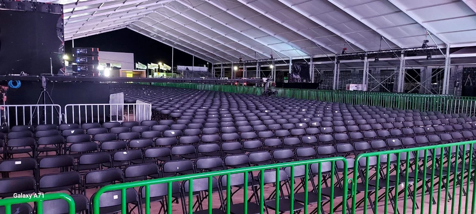 Asientos vacíos en un espacio cubierto para un evento, con escenario y equipo de sonido al fondo, y barricadas verdes en primer plano, en la noche.