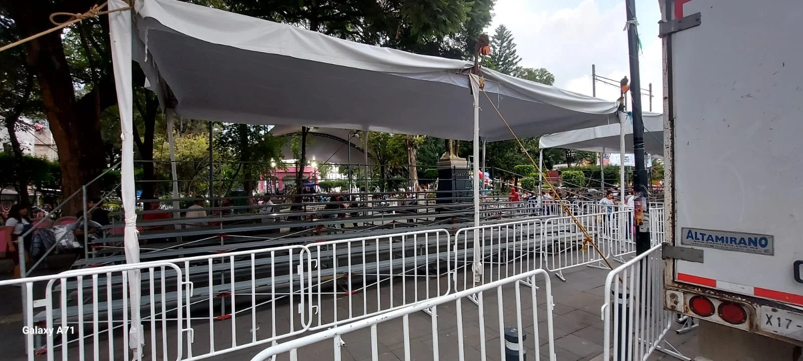 Estructuras metálicas y cercas blancas en un evento público en la calle, con árboles y personas al fondo.