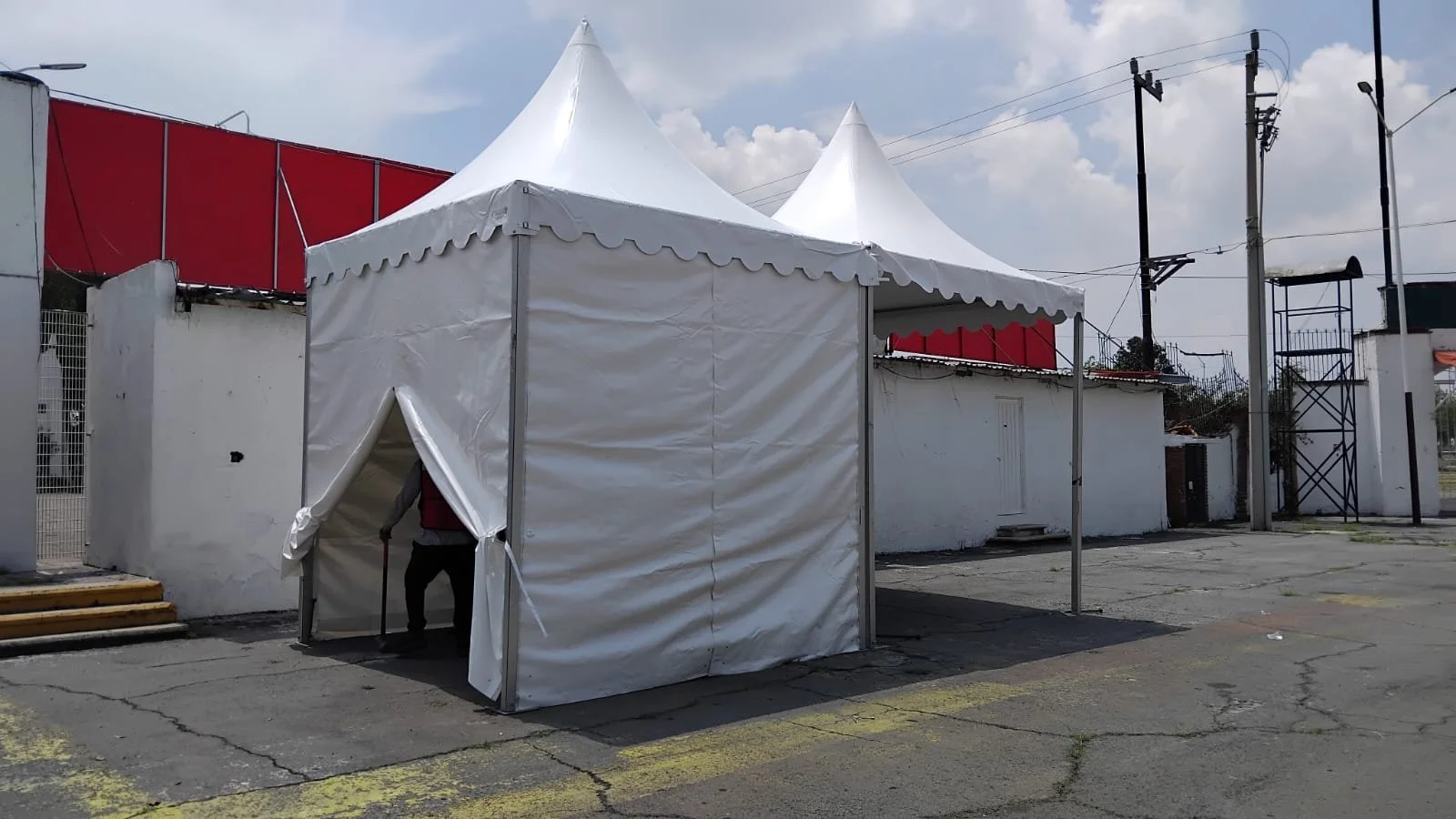 Una carpa blanca en la calle, con una persona entrando, en un entorno urbano con muro blanco, postes de electricidad y cielo nublado.