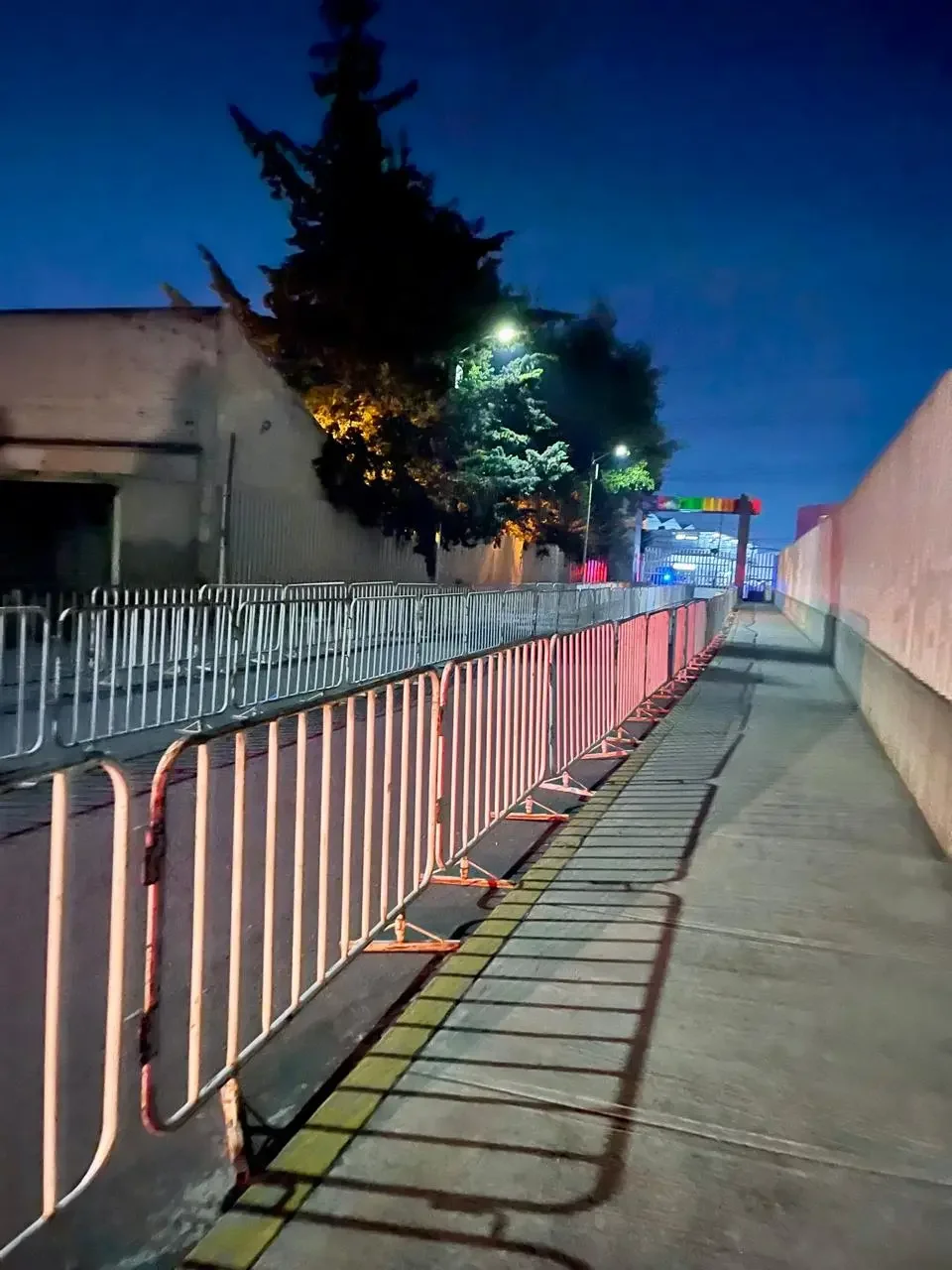 Valla de protección en una acera, con sombra de árboles y postes de luz, en una noche despejada.