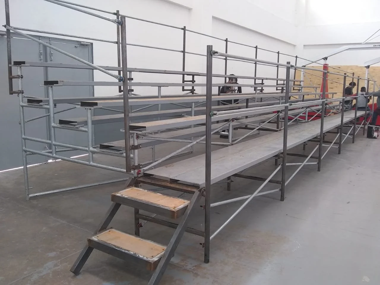 Estructura de andamios de metal y madera en interior, con escaleras y personas en fondo.