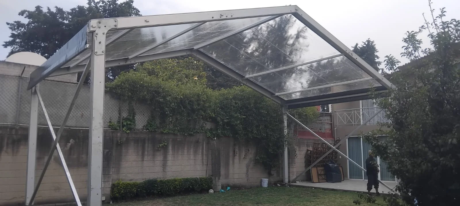 Estructura metálica con techo de acrílico en un jardín, con una persona en la esquina derecha.