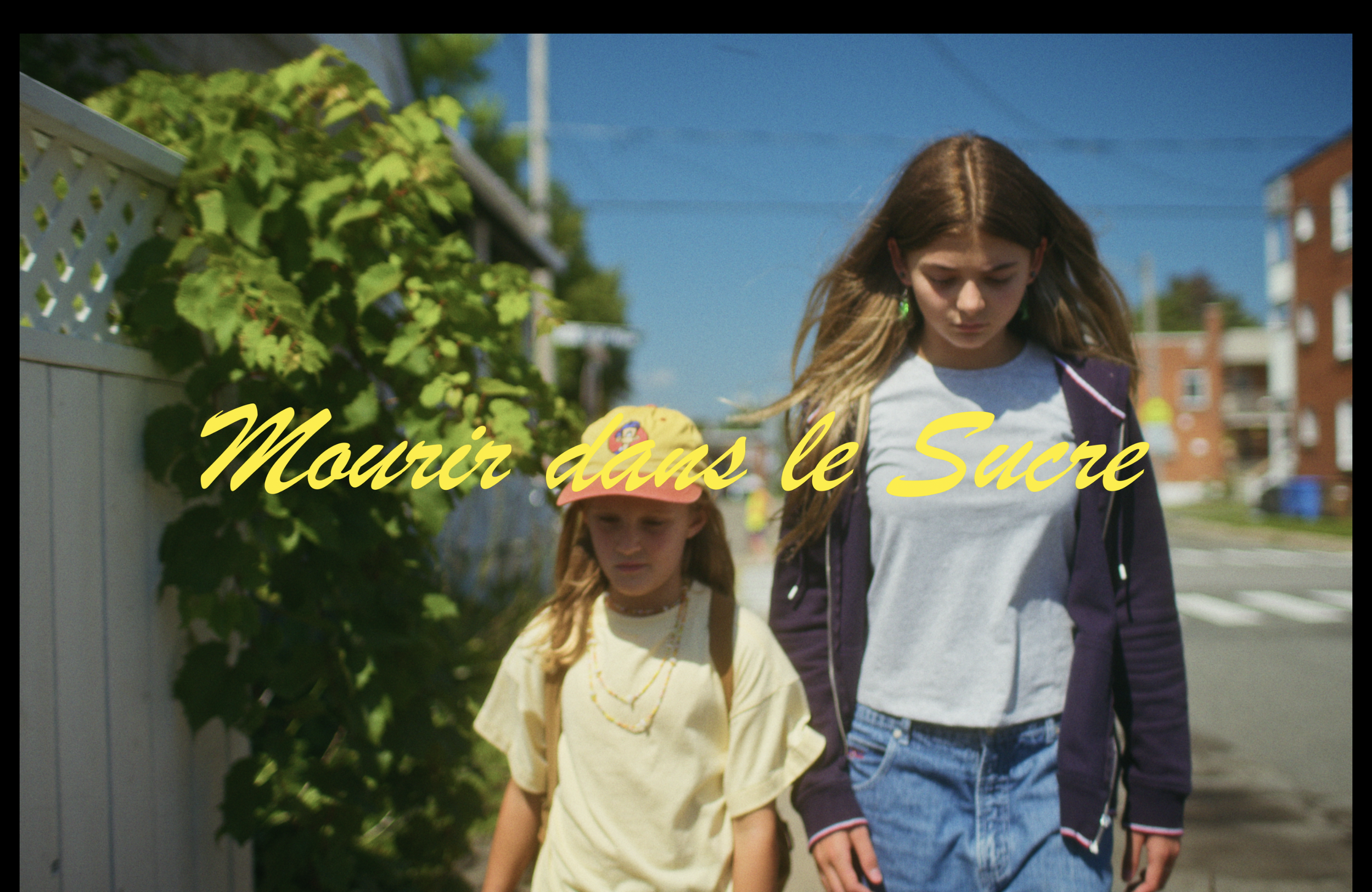Deux filles marchent côte à côte dans une rue ensoleillée, avec des bâtiments résidentiels en arrière-plan, alors que du texte en français "Mourir dans le Suc" est écrit en jaune en bas de l'image.