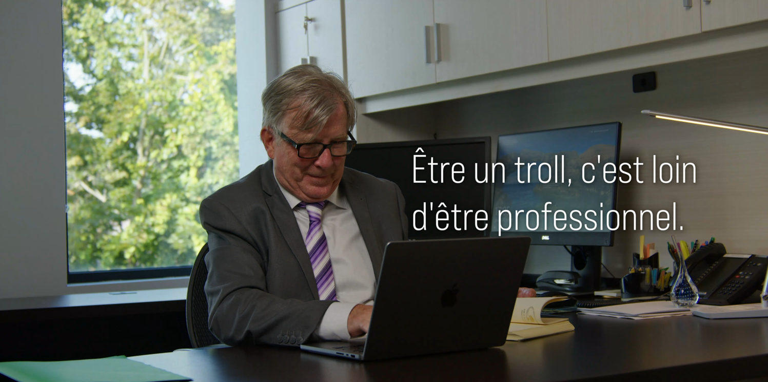 Une personne âgée en costume assise à un bureau avec un ordinateur portable, dans un bureau moderne avec une grande fenêtre laissant entrer la lumière naturelle.