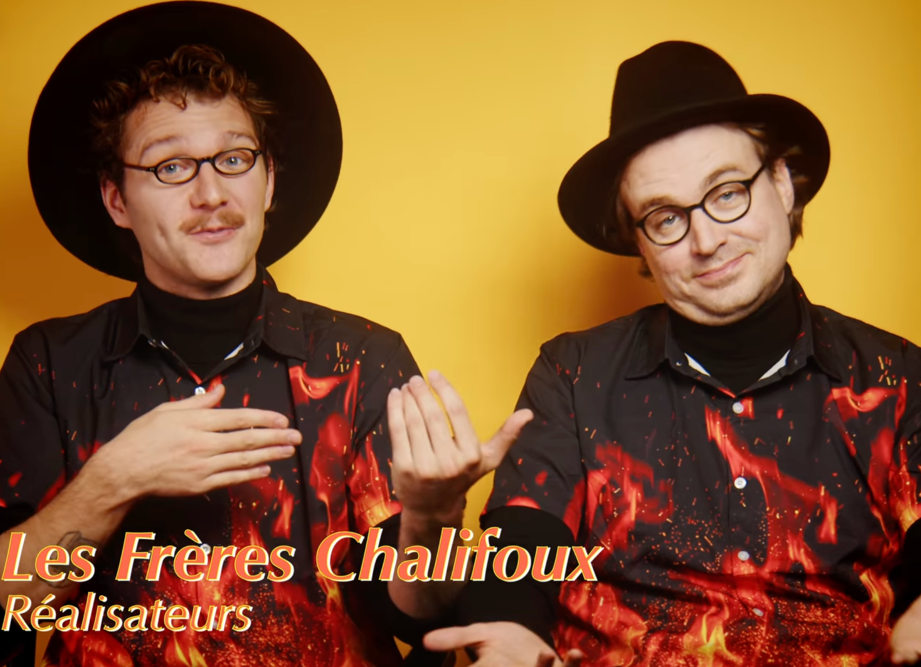Deux hommes portant des chapeaux noirs et des chemises à motif rouge et noir, posant contre un fond jaune. L'un d'eux a une moustache et porte des lunettes, et ils semblent faire une présentation ou une performance.