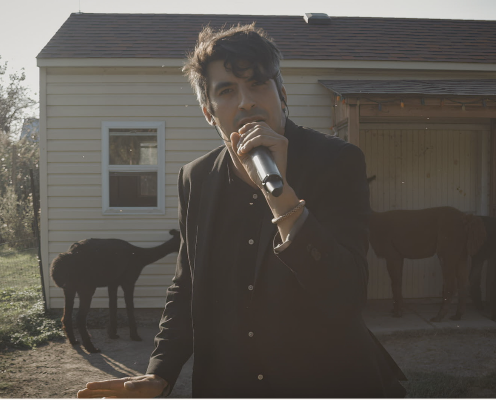 Un homme chante avec un microphone devant une maison avec deux chevaux dans l'arrière-plan.