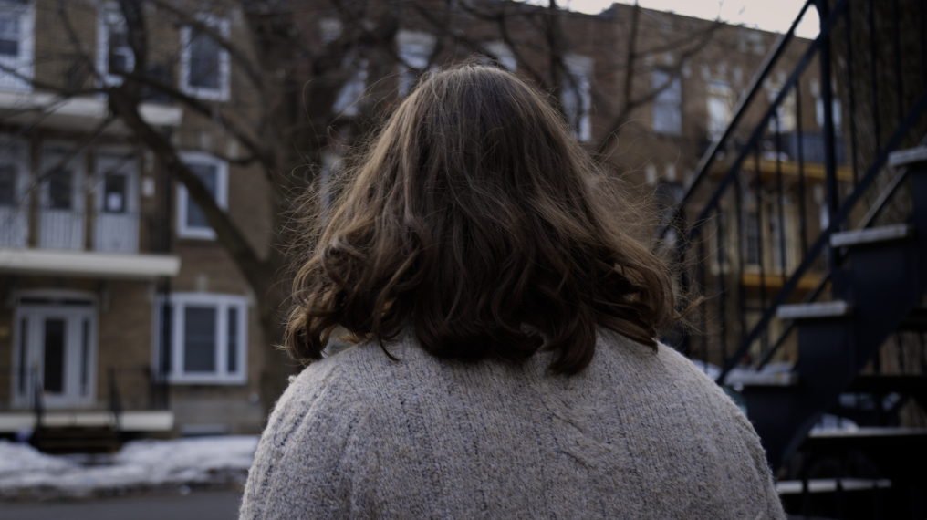 Personne vue de dos, avec des cheveux bruns, portant un pull beige, dans un environnement urbain avec des bâtiments résidentiels et des escaliers en métal.