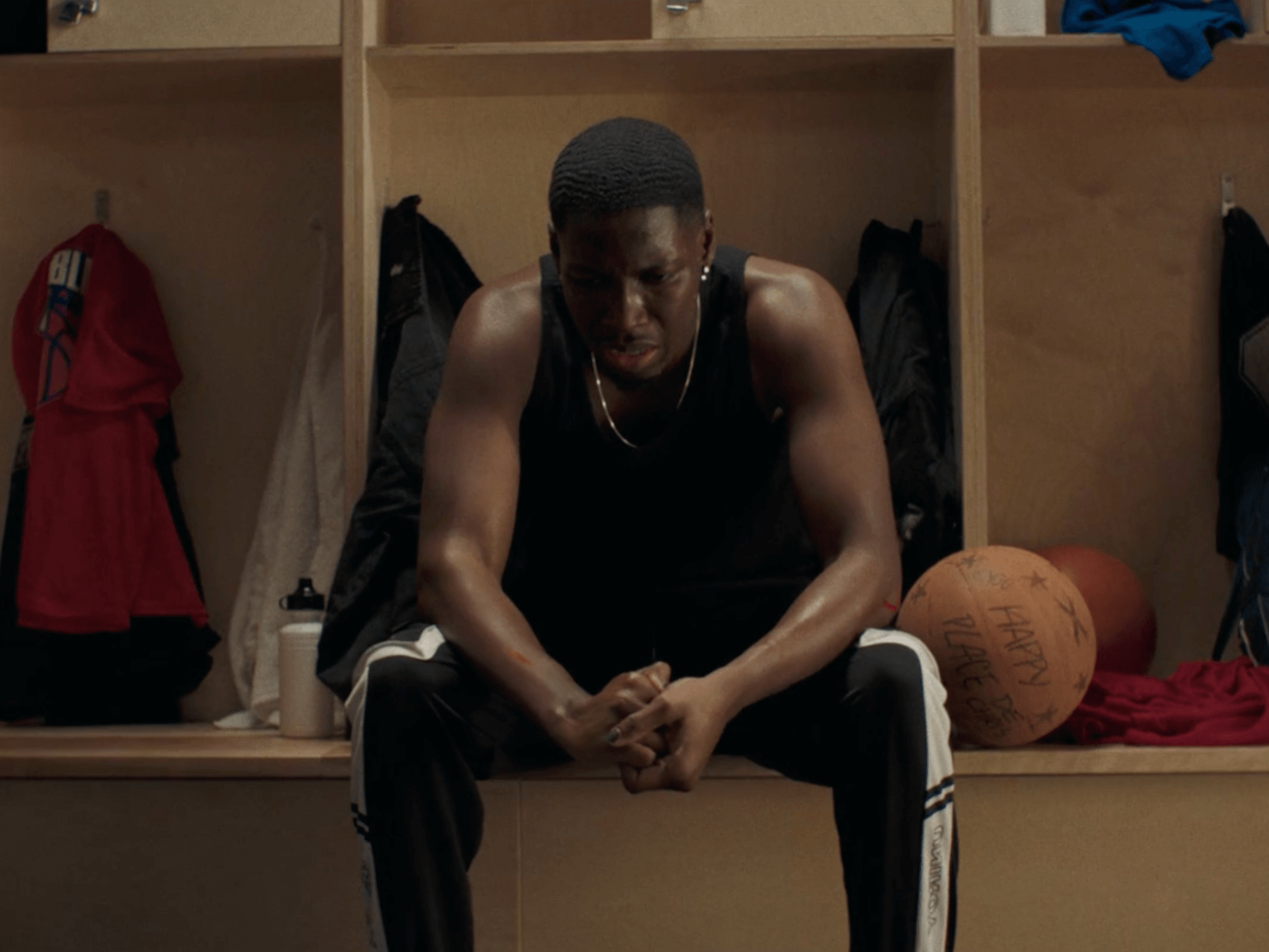 Un jeune homme dans un vestiaire, assis sur un banc, avec une atmosphère de défaite ou de réflexion intense. Il porte un maillot noir, des pantalons de sport, et semble fatigué, le visage exprimant une émotion forte.