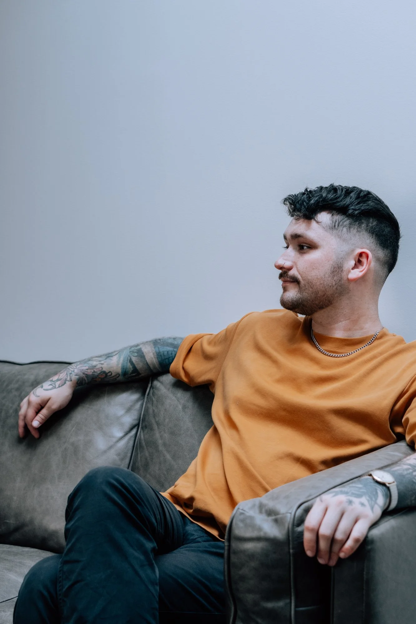 Un homme assis sur un canapé gris, portant un t-shirt orange, avec des tatouages sur le bras, regarde vers la droite.