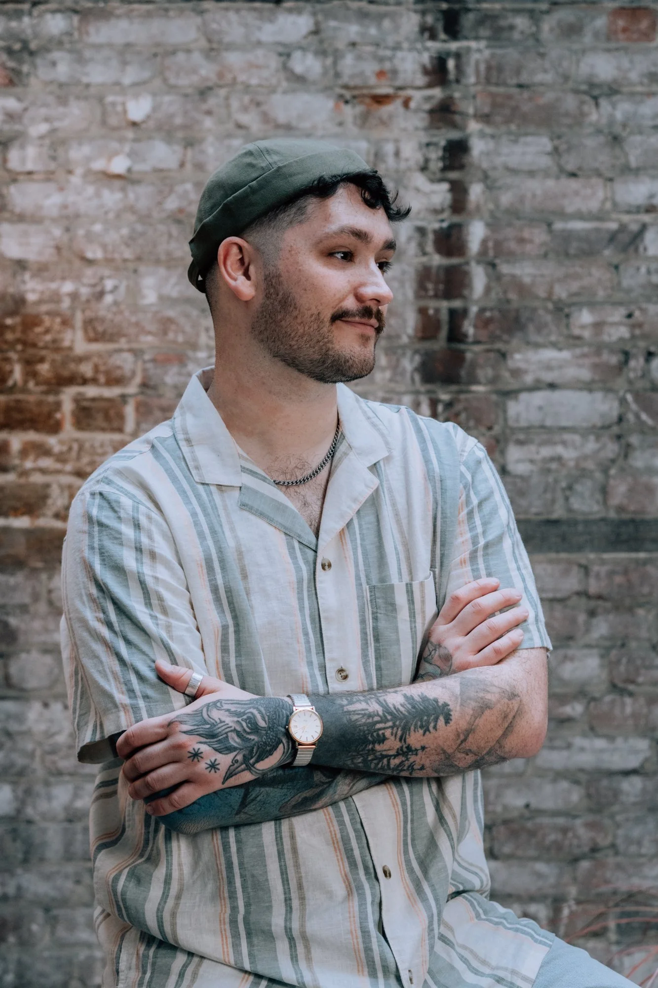 Un homme debout avec les bras croisés, portant une chemise à rayures, une montre et un tatouage sur le bras, avec un mur en briques en arrière-plan.