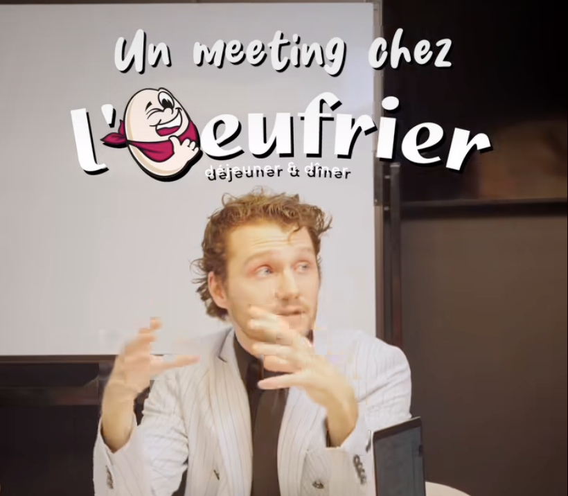 Photo d'un homme en train de parler lors d'une réunion chez l'épicier, avec un panneau en arrière-plan indiquant 'Un meeting chez l'épicier', avec une illustration d'un œuf en smiley