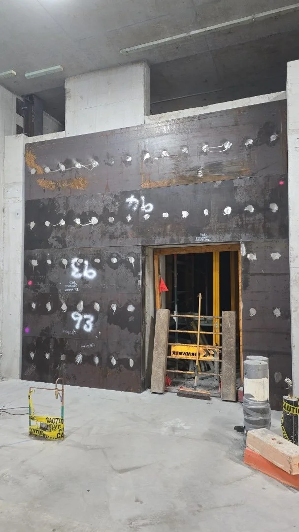 Construction site showing an unfinished wall with drilled holes, spray marks, and markings, with caution tape and construction materials on the floor.