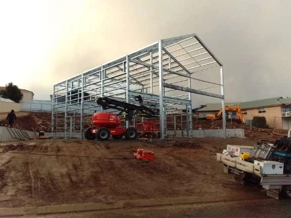 Steel framework of a building under construction with construction equipment on site, dirt ground, and neighboring buildings in the background.