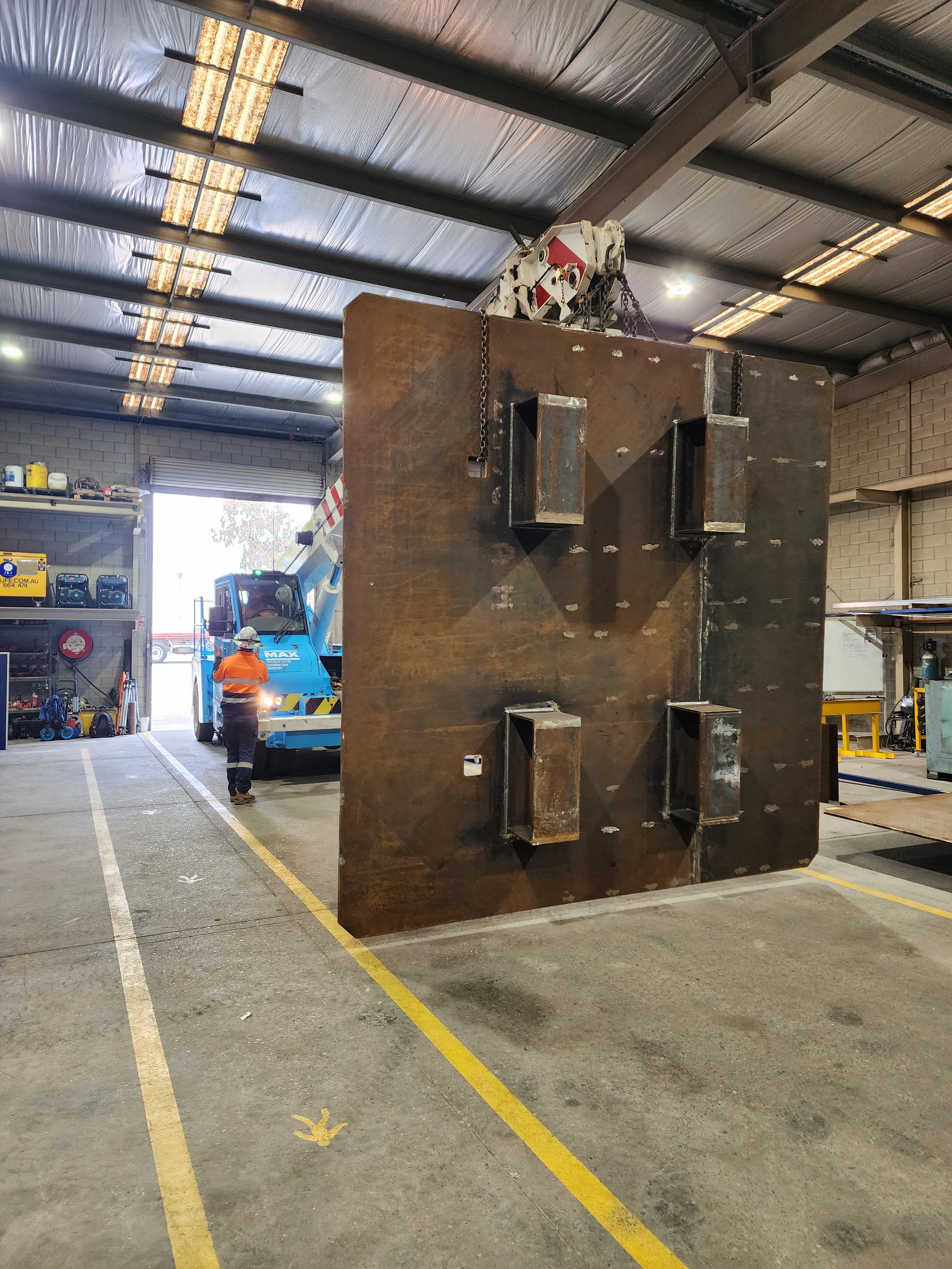 Large steel plate with four mounting brackets inside an industrial warehouse, with a worker and crane in the background.