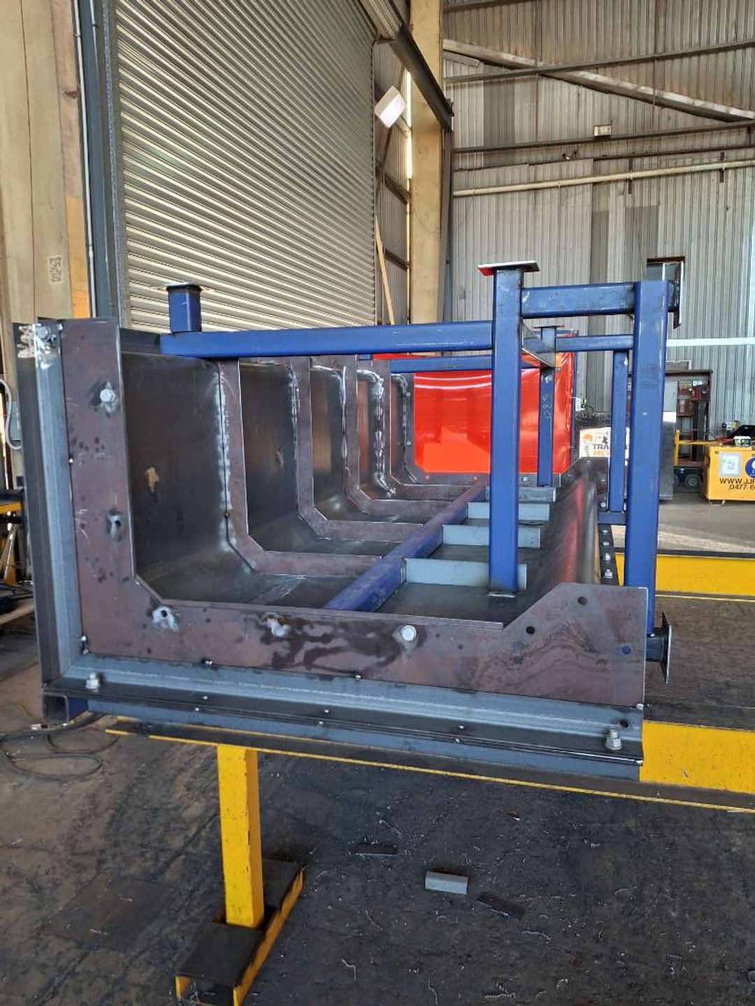 Industrial metal structure inside a warehouse with steel beams and a partially assembled frame, possibly for a machine or equipment, with a yellow support stand underneath.