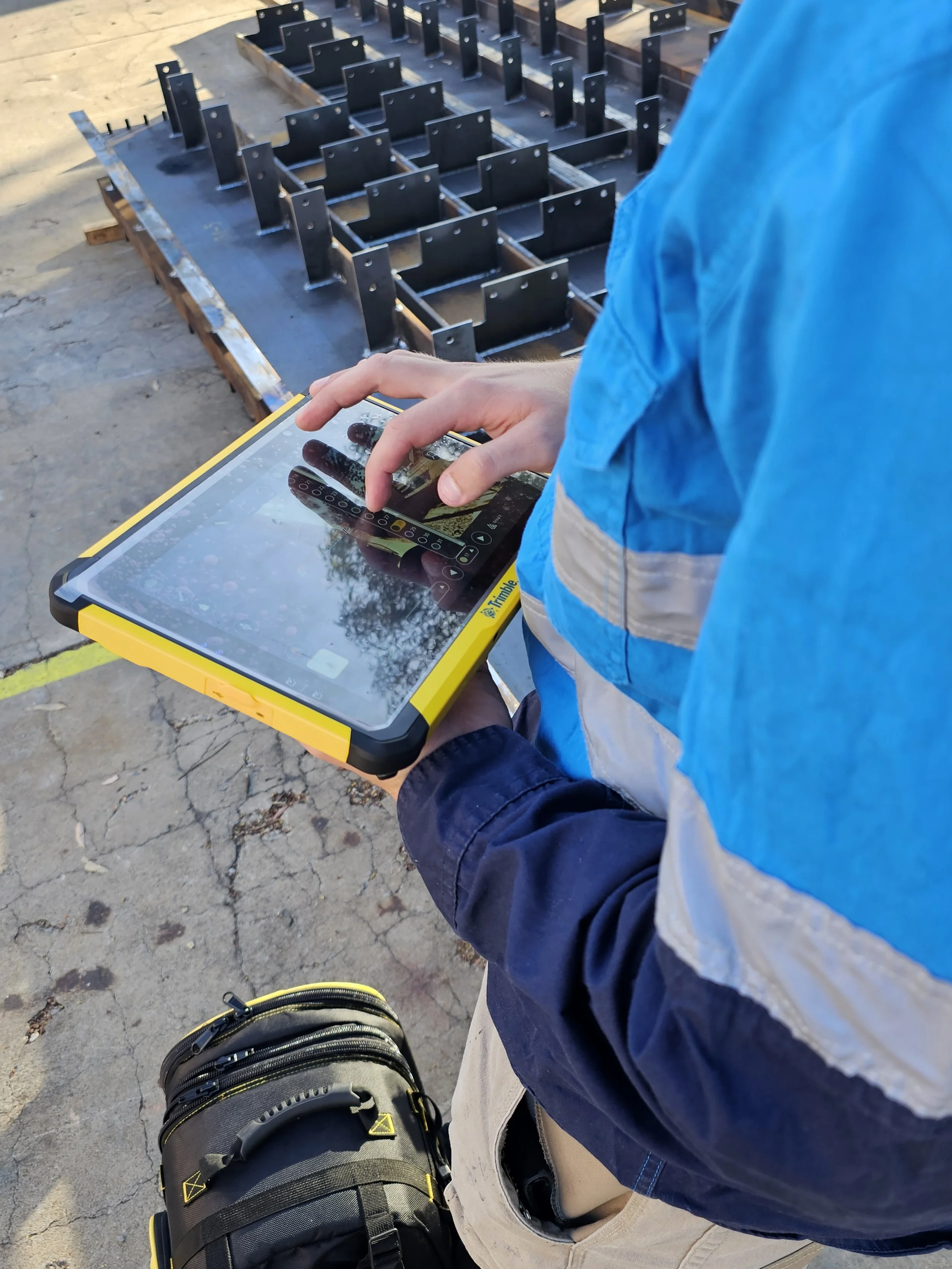 A person holding a rugged tablet with a stylus, working outdoors at a construction or industrial site. In the background, there are metal brackets or frames mounted on a wooden pallet.