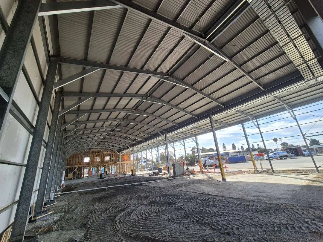 Under-construction building with metal roof and open sides, construction workers in the background, dirt ground, and construction materials around.