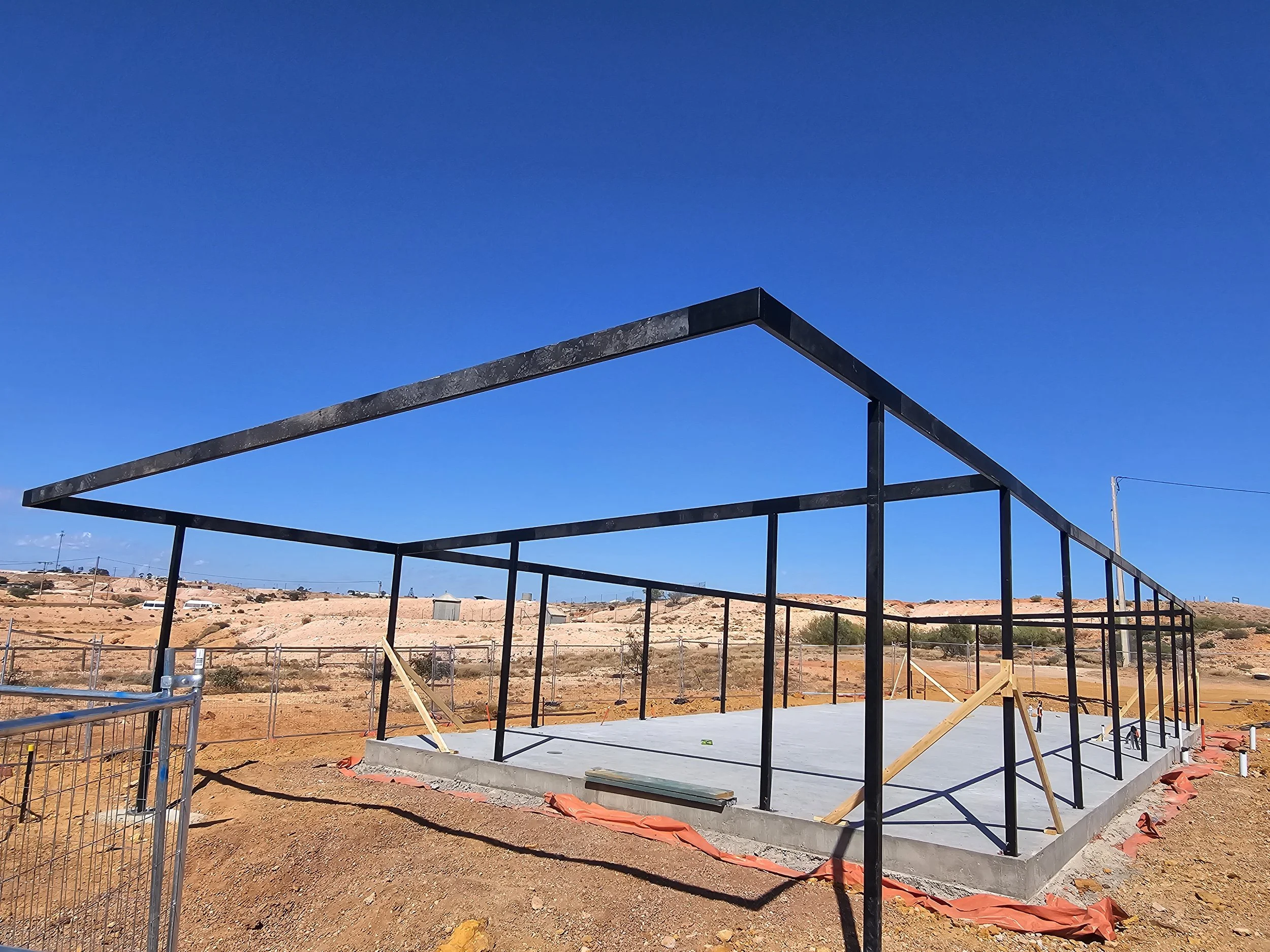Construction site with a black steel frame structure over a concrete slab in a desert landscape under a clear blue sky.