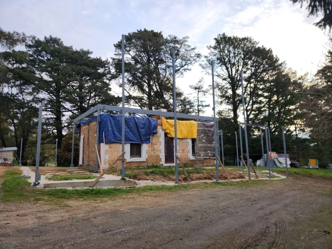 House under construction with metal framing and temporary coveralls, surrounded by trees.
