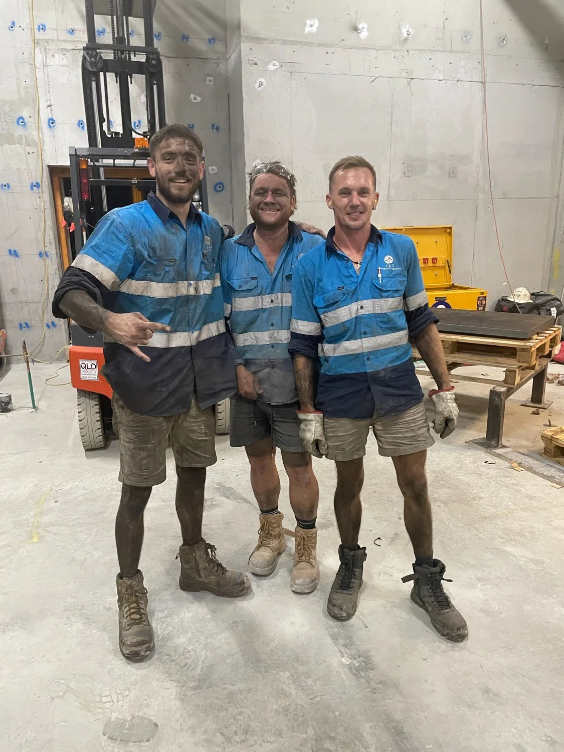 Three construction workers wearing blue shirts and work gloves, standing together inside a construction site with muddy shoes and smiling.